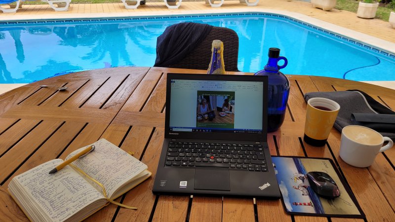 Aufgeklapptes Laptop auf Terrassentisch am Pool, daneben ein Notizbuch und Getränke