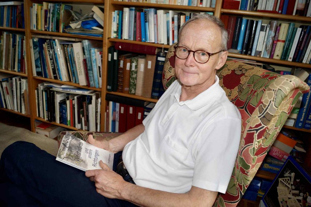 Andreas Hufschmidt in weißem Poloshirt in Sessel vor Bücherregal, in der Hand das Buch Westwärts